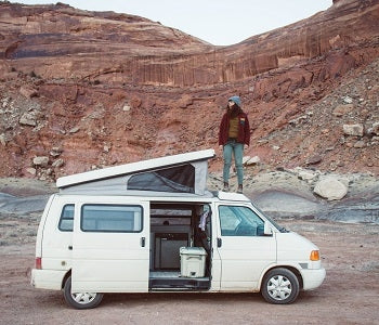 Pictured: Woman standing on top of white camper van with rocky terrain in background