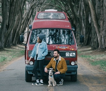 Pictured: a couple and their dog pose in front of a red camper van on a road with a canopy of trees above