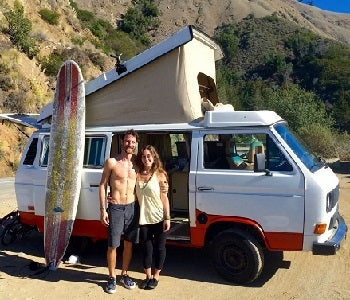 Pictured: couple stands next with surfboard in front of white camper van on the beach
