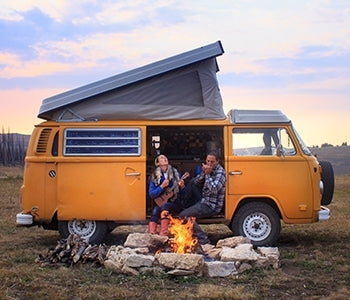 Pictured: couple sitting in the open door of a yellow camper van with a campfire burning in front of them