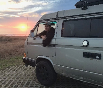 Pictured: man leaning out the open window of a parked camper van with the sun setting in the background