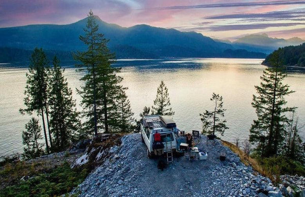 Camper van parked by scenic lake and mountains at sunset, outdoor camping setup visible.