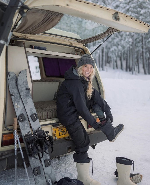 Woman in winter gear sitting in a camper van with skis, snow boots, and snowy forest background
