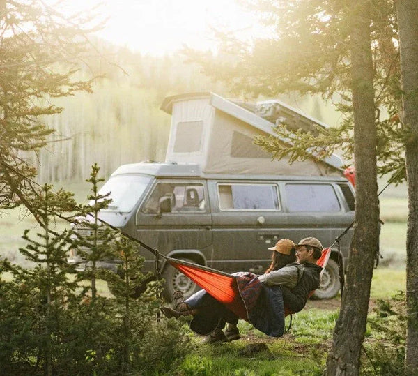 Couple relaxing in hammock near camper van in forest, outdoor camping adventure scene