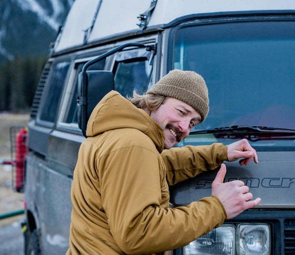 Man in brown jacket and beanie giving thumbs up next to a Syncro camper van outdoors