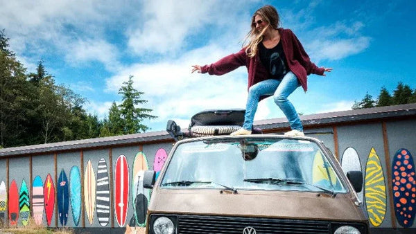 Woman posing on top of a vintage van in front of a colorful surfboard mural outdoors