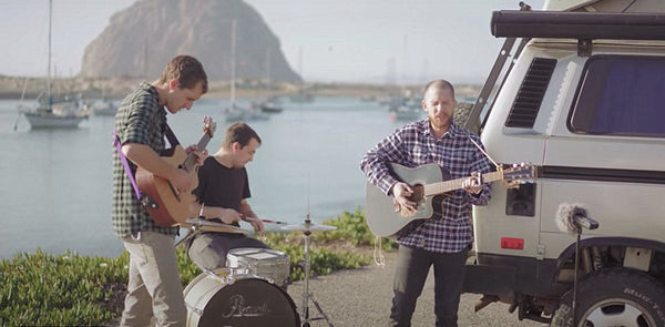 Outdoor band playing guitar and drums by a camper van near water and boats