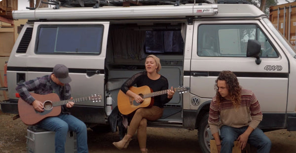 Pictured: three people, two with guitars, sitting in front of a silver van