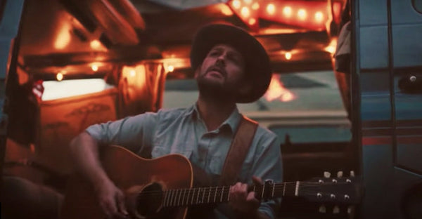 Man playing acoustic guitar beside camper van with string lights at dusk