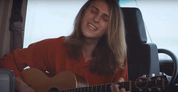 Person with long hair playing acoustic guitar and singing inside camper van