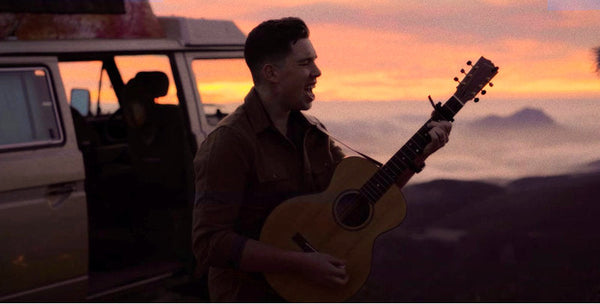 Man playing acoustic guitar by a camper van at sunset in scenic outdoor setting