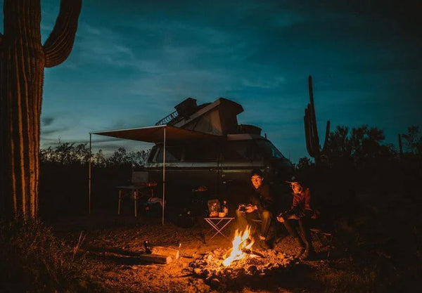 Two people camping by a van with rooftop tent, sitting around a campfire at dusk in the desert.