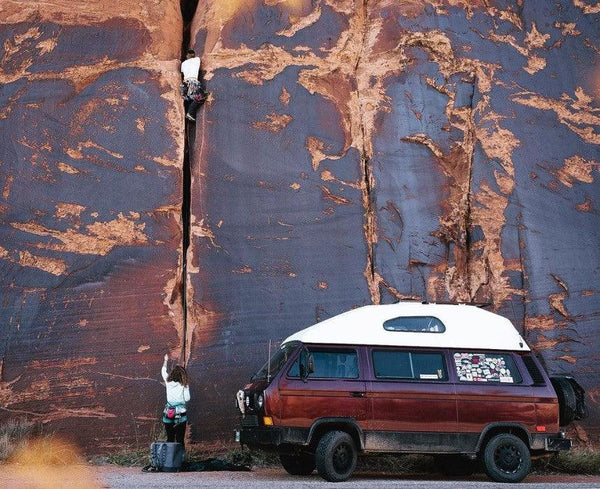 Vanagon camper van parked by red rock canyon, person rock climbing crack with belayer