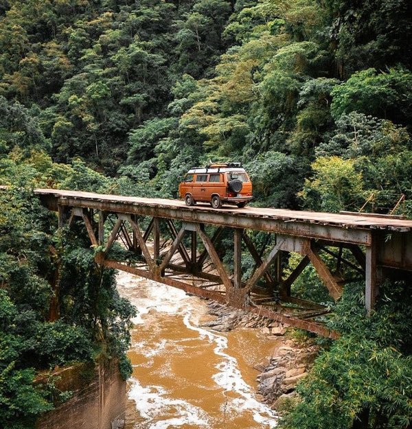 Orange vintage van crossing a narrow bridge over a river in a lush green forest