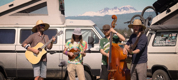 Four musicians playing acoustic instruments beside camper vans with mountains in the background