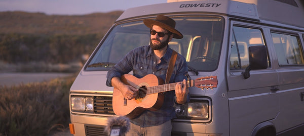 Man playing acoustic guitar by camper van at sunset, wearing hat and sunglasses