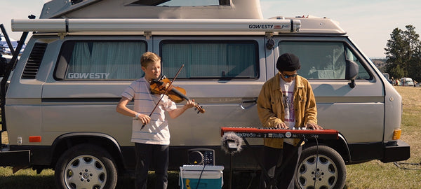 Two musicians performing outdoors beside a silver camper van, one playing violin, one on keyboard.