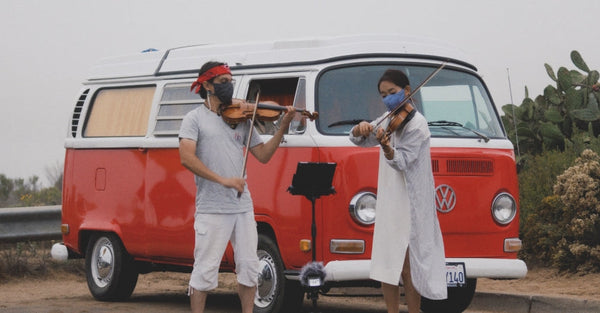 Two people playing violins in front of a red Volkswagen camper van outdoors