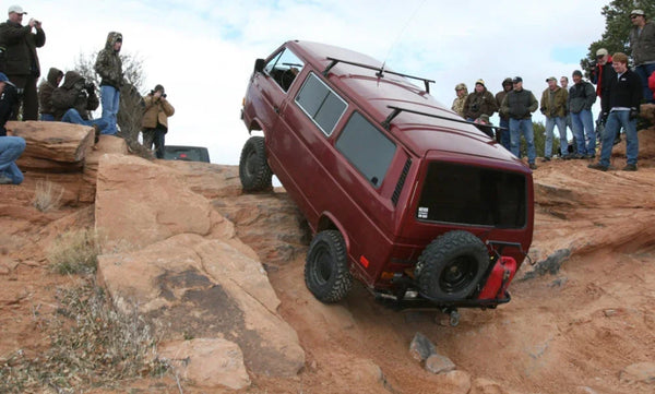 Red VW camper van off-roading on rocky terrain surrounded by spectators