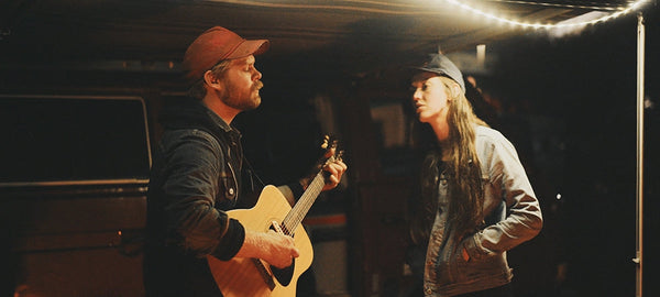 Two people by a van at night, one playing guitar, under string lights, camping scene.