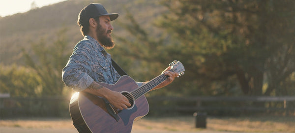 Man playing acoustic guitar outdoors in sunlight, casual outfit, nature background