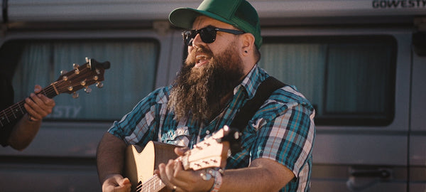 Bearded man in sunglasses and cap playing acoustic guitar outdoors by camper van
