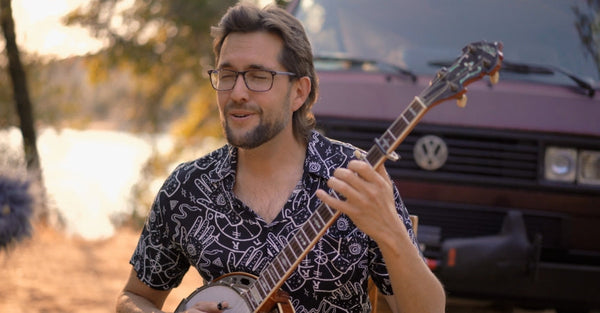 Man playing banjo outdoors in front of a maroon Volkswagen van camper