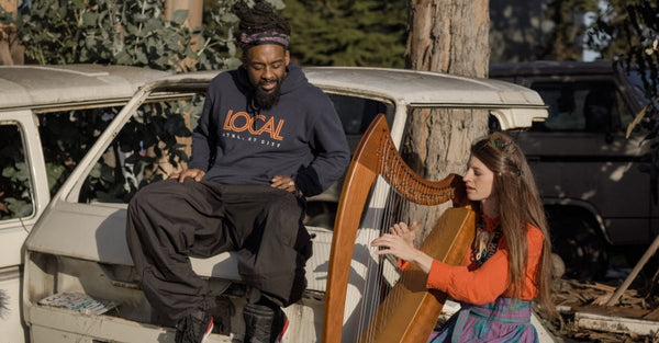 Man in LOCAL sweatshirt sitting on vintage van, woman playing harp outdoors