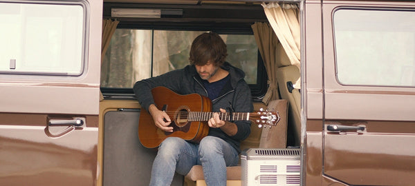 Man playing acoustic guitar inside a brown camper van with open side door and beige curtains.