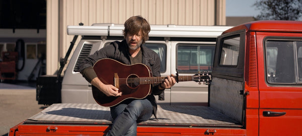 Man playing acoustic guitar sitting on red Volkswagen truck bed, camper vans in background