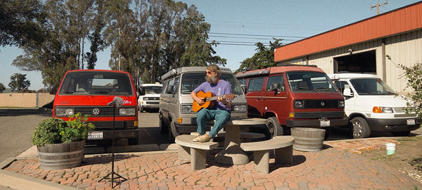 Man playing guitar on stone picnic table in front of parked Volkswagen camper vans