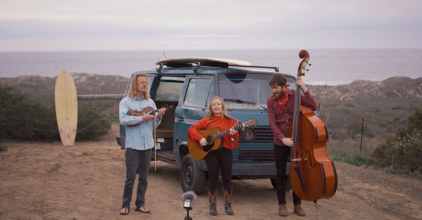 Outdoor band performance by a camper van with surfboard, ocean cliffs in background