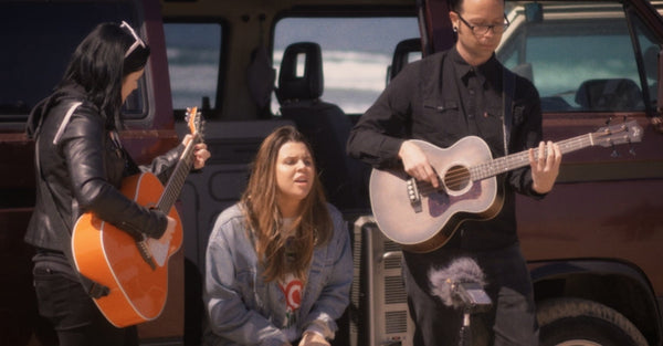 Three musicians performing with acoustic guitars by a van at the beach during daylight.