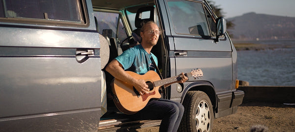 Man playing guitar beside classic camper van by a scenic waterfront outdoors