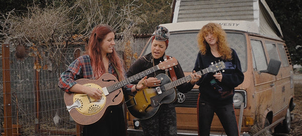 Three women playing guitars outdoors in front of a vintage Volkswagen camper van with pop-top.