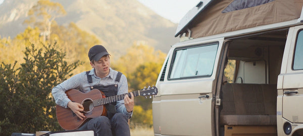 Man playing acoustic guitar outdoors beside a vintage camper van with pop-top roof in nature.