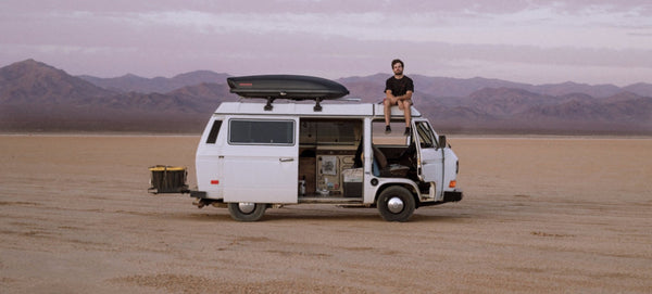 White camper van parked on desert plain with mountains in background, man sitting on roof