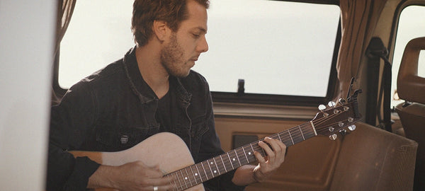 Man playing acoustic guitar inside a camper van with sunlight streaming through windows
