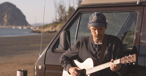 Pictured: Jack Symes with a guitar leaning on the side of a camper van with the beach in the background