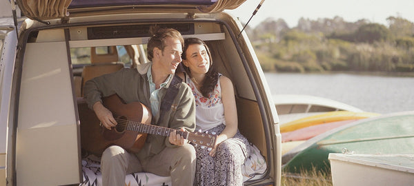Couple sitting in camper van by a lake, man playing guitar, enjoying outdoor adventure.
